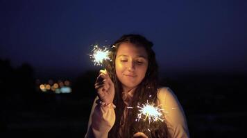 uma lindo jovem menina com grandes Sombrio cabelo carrinhos contra a pano de fundo do a cidade às noite e detém fogos de artifício dentro uma Boa humor. lento movimento. 4k video