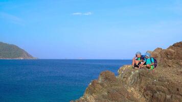 Rapazes com mochilas sentado em uma Rocha em a margens do a Mediterrâneo mar. crianças descansar e lançar pedras para dentro a água. a conceito do aventura e Novo descobertas. video
