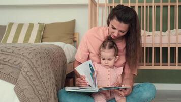 puericultura às lar, criança proteção, Novo vida, lazer com bebê. mãe lê uma livro para a criança enquanto sentado perto a cama às casa video