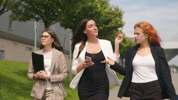 Business ladies walking in the park with mobile gadgets in their hands smiling and talking. Business meeting of three women on the background of a futuristic building in the park photo