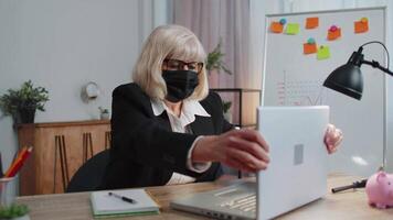 Senior employee wears face mask works alone from home office sits at desk using laptop computer video