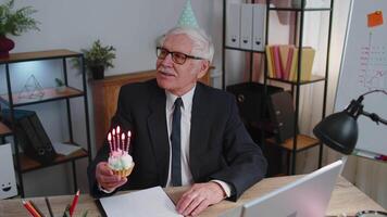 Senior old business man celebrating birthday in office holding small cake with candles making a wish video