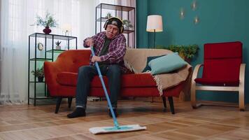 Happy senior grandfather housekeeper is mopping floor in living home room, listening music, dancing video