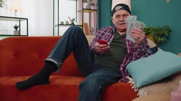 sorridente Senior homem segurando dinheiro e Smartphone animado sobre Móvel aplicativo esporte aposta licitação ganhar às casa video
