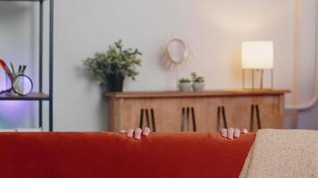 Man playing hide and seek peekaboo game hiding behind sofa looking at camera, positive smiling video