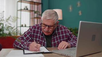 Senior man working on laptop computer, making notes on sheet of paper, upset by poor bad results video