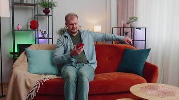 heiter jung Mann Sitzung auf Sofa, mit Handy, Mobiltelefon Telefon Teilen Mitteilungen auf Sozial Medien Anwendung video