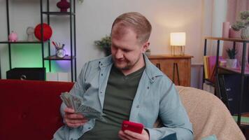 sorridente feliz jovem homem contando dinheiro dinheiro e usar Móvel telefone calcular doméstico contas às casa video