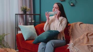 Young Caucasian sick woman wearing medical protection mask sitting in living room looking at camera video