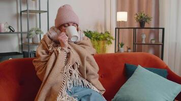 Young sick woman wear hat wrapped in plaid sit alone shivering from cold on sofa drinking hot tea video