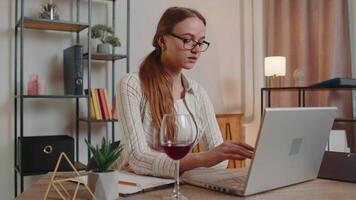 focalizzata donna libero professionista potabile vino mentre Lavorando digitando in linea su il computer portatile a casa distanza ufficio video