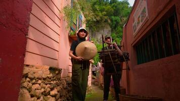 Two men walking in a narrow alley, one carrying a large hat and the other carrying wooden items, with rustic walls on either side. video