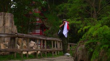 Traveler with a backpack standing on a wooden bridge in a lush park, admiring a traditional red pagoda. video