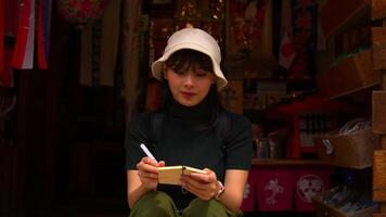 Smiling woman in a hat writing in a notebook while sitting on wooden steps with colorful flags in the background. video