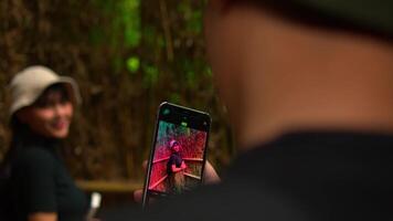 Over-the-shoulder view of a person taking a photo of a smiling woman with a smartphone in a natural setting. video