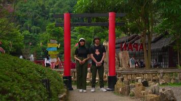 Tourist couple posing under a traditional red torii gate at a historic site with lush greenery in the background. video