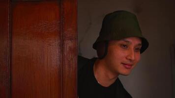 Portrait of a young man in a green bucket hat, smiling subtly indoors with a warm, dimly lit background. video