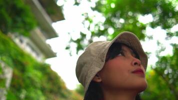 Woman in hat and scarf looking upwards with blurred greenery and ladder in the background. video