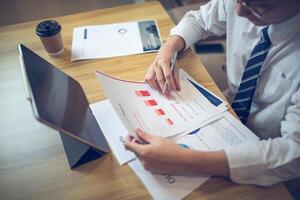 Businessman analyzing bar graph data with pen in hand, laptop and coffee on desk. Business concept photo