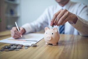 persona en un negocio camisa ahorro dinero en un cerdito banco, con monedas y financiero informes en el mesa. ahorro dinero negocio concepto foto