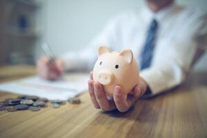 persona en un negocio camisa ahorro dinero en un cerdito banco, con monedas y financiero informes en el mesa. ahorro dinero negocio concepto foto