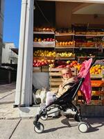 Budva, Montenegro - 18 october 2023. Little girl sitting in a stroller near the fruit stand photo