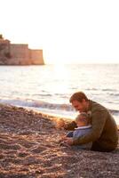 Smiling dad pouring pebbles into the palm of a little girl sitting on the beach by the sea photo