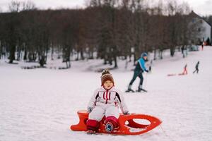 pequeño niña se sienta en un trineo en un Nevado llanura en contra el fondo de trineos y esquiadores foto