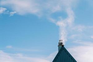 Smoke billows above the chimney of a triangular green corrugated roof of a cottage photo
