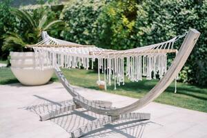 White wicker hammock with tassels hangs on a wooden stand in a green garden photo