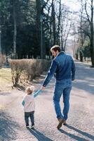 papá con un pequeño niña caminar participación manos a lo largo el la carretera en el primavera parque. espalda ver foto