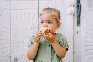 Little girl gnaws a red apple while standing near a white carved wooden door and looks away photo