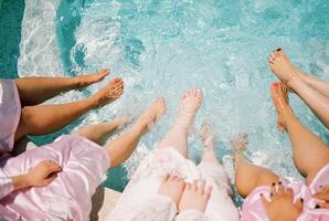 novia y su damas de honor sentar en el borde de el piscina con su pies en el turquesa agua. recortado sin rostro foto