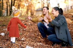 Happy mom and dad sprinkle dry leaves on a little girl sitting in the autumn forest photo