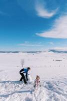 pequeño niña y su papá son haciendo un monigote de nieve por laminación grande bolas de nieve a través de el césped foto