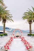 Rows of chairs line a white path decorated with red flowers in front of a round wedding arch on the pier photo