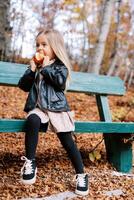 pequeño niña roe un manzana mientras sentado oblicuo en un de madera banco en el otoño bosque foto