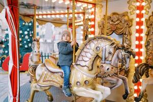 pequeño niña paseos un juguete caballo en un carrusel en el cuadrado cerca un decorado Navidad árbol foto