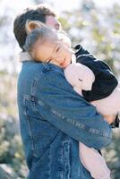 Little smiling girl put her head on her dad shoulder while sitting in his arms in the park. Side view photo