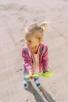 Little girl with a toy shovel in her hands stands on the sand and looks away. Top view photo