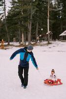 Dad carries a small child on a sleigh along the snowy edge of the forest, looking back photo