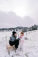 sonriente madre se sienta en un de madera trineo y mira a un pequeño niña en pie en un Nevado colina cerca foto