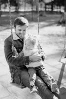 Smiling dad with a little girl in his arms sits on a chain swing. Black and white photo