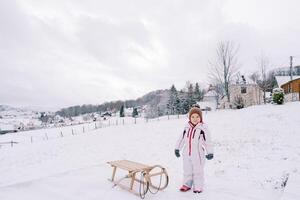 Little smiling girl stands near a wooden sled on the edge of the village photo