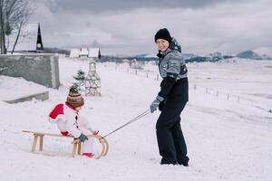 Smiling mother pushing a wooden sled with a little girl down a snowy hill photo