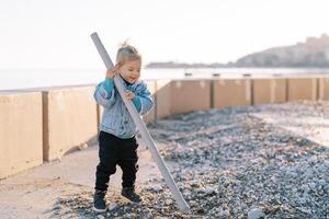 Little laughing girl picking pebbles with a plastic pipe while standing near the fence on the seashore photo