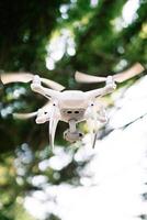 White four-engine drone hovers in the air in a green park photo