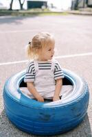 pequeño niña se sienta en un grande azul neumático en el patio de recreo y mira lejos foto