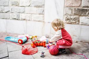 Little girl is playing with different colorful toys while squatting against a stone wall photo