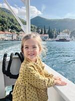 Little girl stands leaning on the side of a boat floating on the sea towards a mountainous shore photo